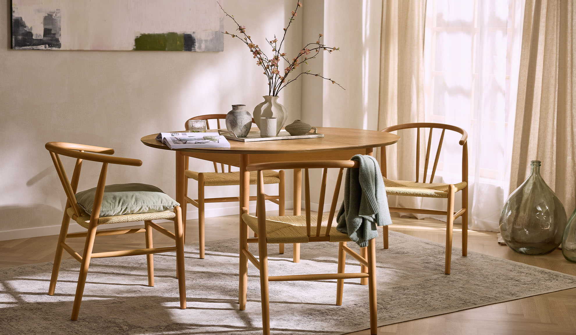 Svetla trpezarija sa okruglim drvenim stolom, drvenim stolicama sa sedištima od užeta i cvetnim dekorom.