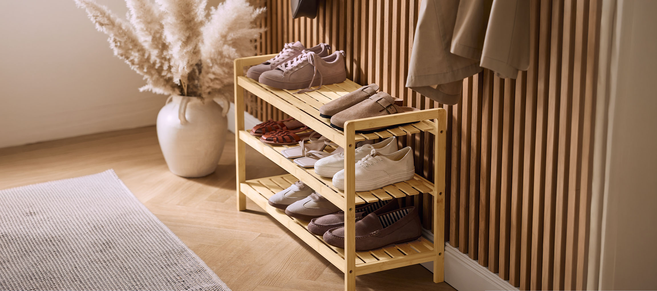 Drveni stalak za cipele sa raznim cipelama, vazom sa pampas travom i kaputom na zidu.