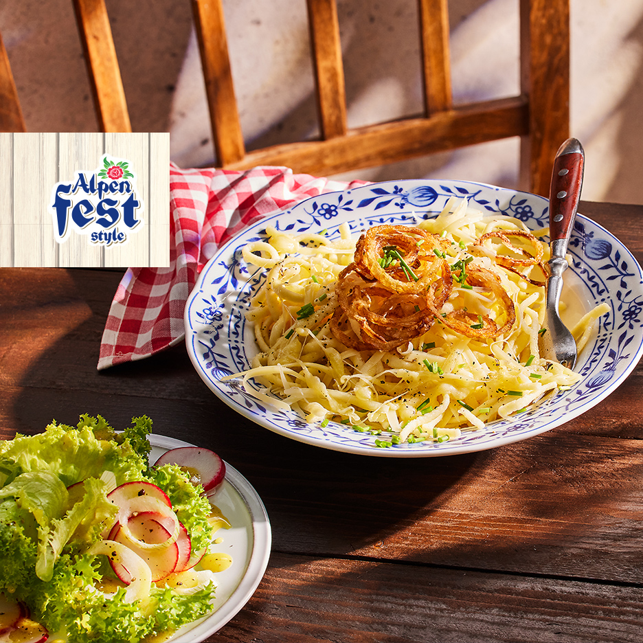 Tanjir špecle sa prženim lukom i salata sa rotkvicama na drvenom stolu, sa logom „Alpen fest style“.