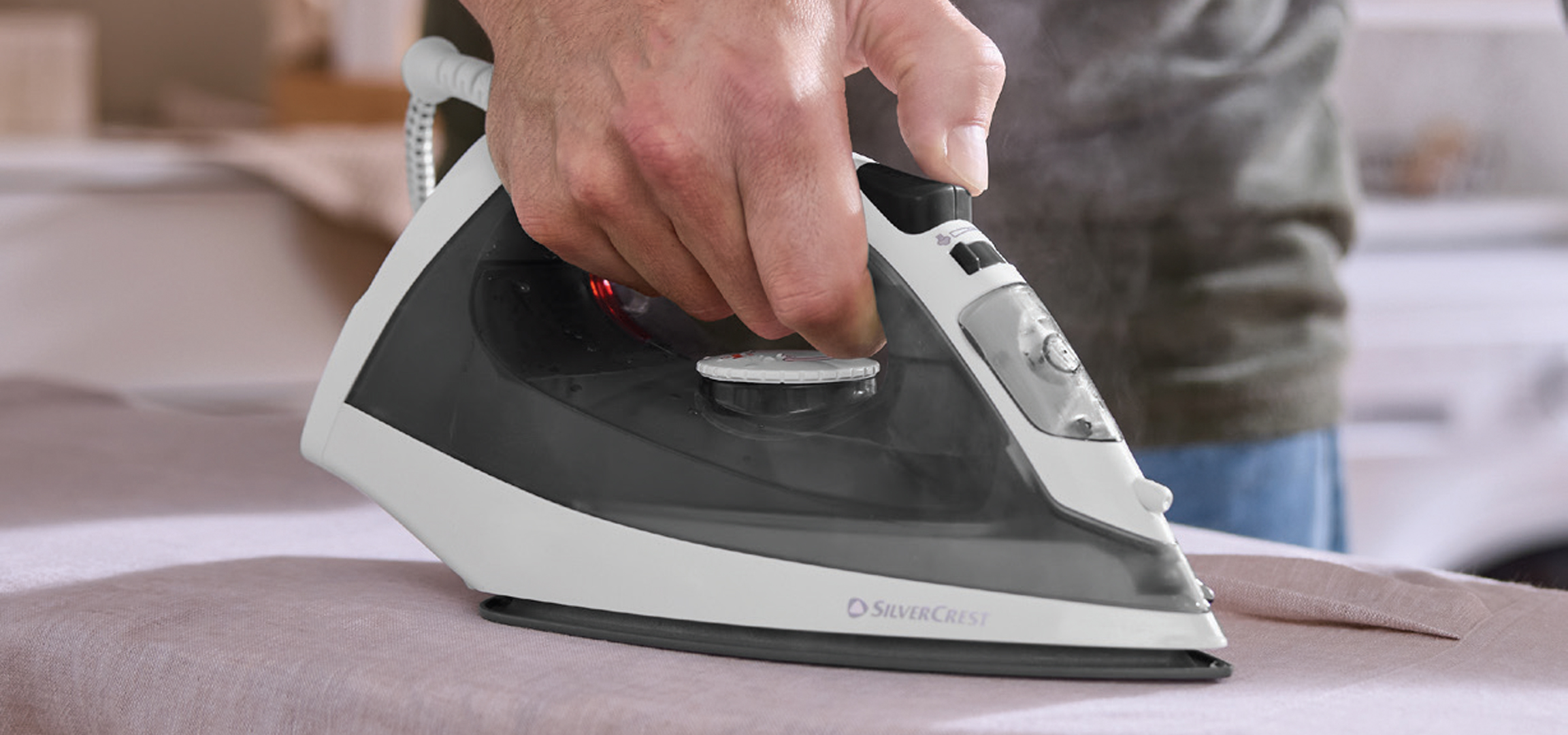 Osoba pegla veš SilverCrest peglom, pored su korpe za veš i SilverCrest ručni aparat za peglanje na paru.