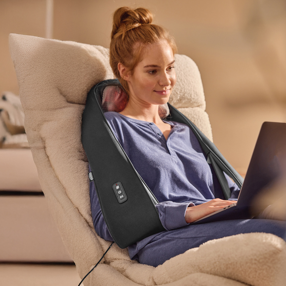 Žena sa masažerom za vrat i ramenima, opušteno radi na laptopu u fotelji.