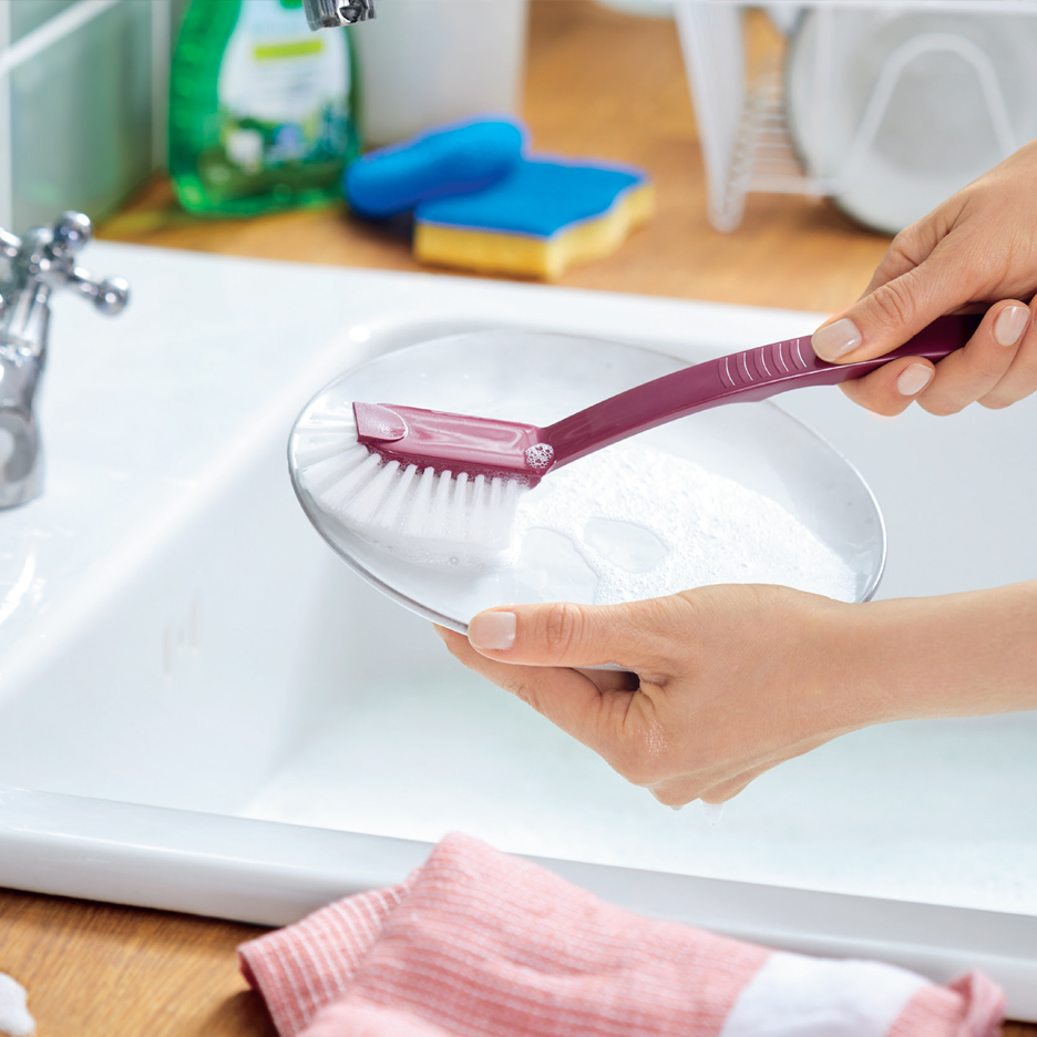 Žena pere tanjir četkom za sudove u sudoperi, sa deterdžentom i sunđerima u pozadini.