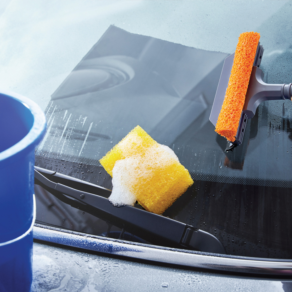 Pranje vetrobranskog stakla automobila sunđerom i brisačem, sa kofom u pozadini.