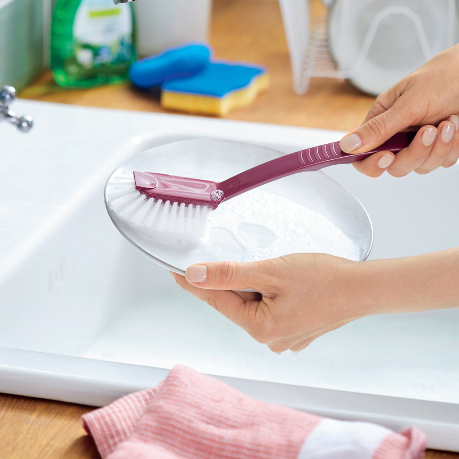 Ruke peru beli tanjir ljubičastom četkom za sudove u kuhinjskoj sudoperi.