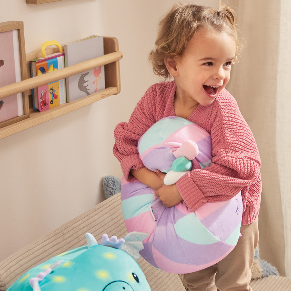 Devojčica grli šareni plišani jastuk, smeje se, pored police sa knjigama.