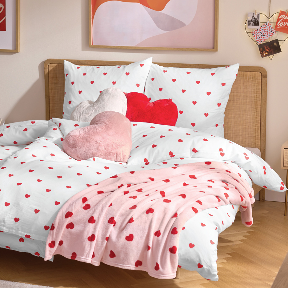Krevet sa belom posteljinom sa crvenim srcima, ružičastim ćebetom i jastucima u obliku srca.