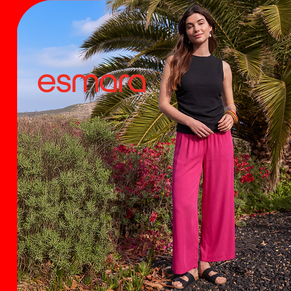 Žena u crnoj majici bez rukava i širokim roze pantalonama, stoji u prirodi sa palmama.