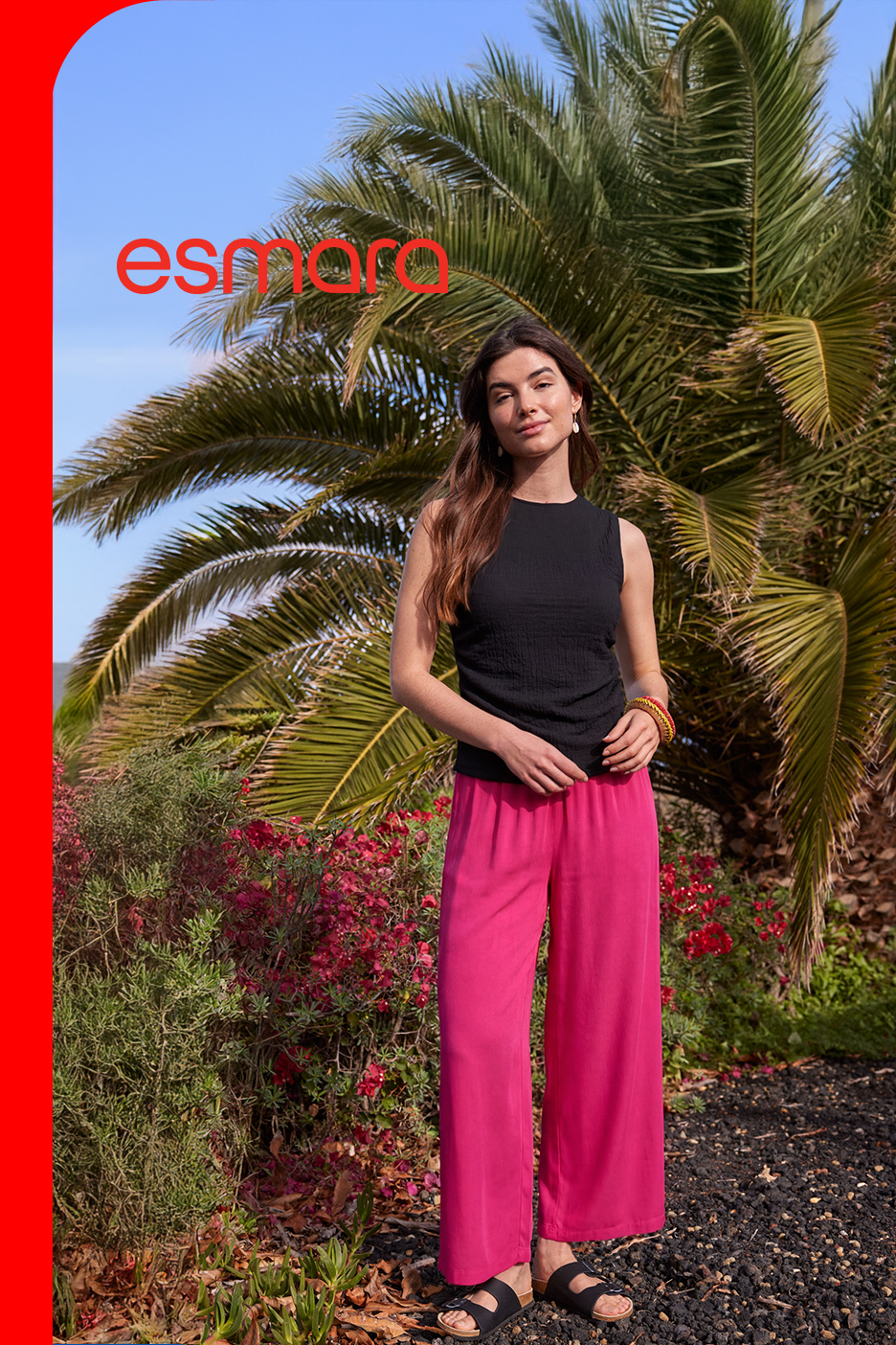 Žena u crnoj majici bez rukava i širokim roze pantalonama, stoji ispred palmi.