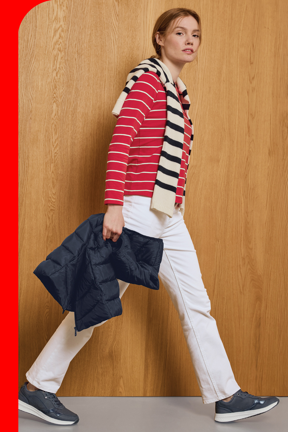 Žena u crvenoj prugastoj majici, belim pantalonama i tamnoplavim patikama, drži tamnoplavu jaknu.