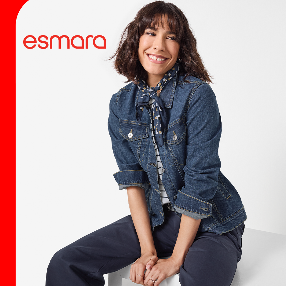 Žena u teksas jakni, prugastoj majici i tamnim pantalonama, sa maramom oko vrata, sedi i smeje se.