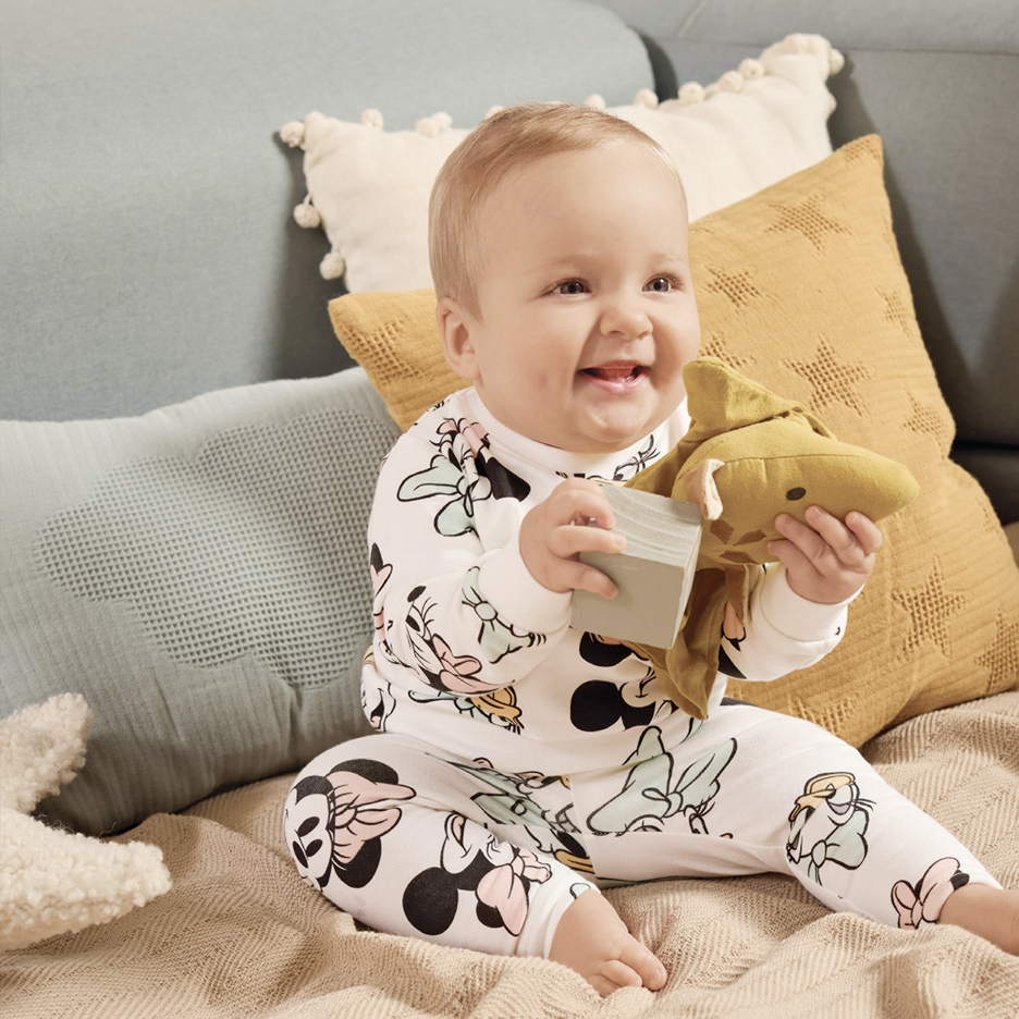 Beba u beloj pidžami sa dezenom sedi na kauču i drži igračke.