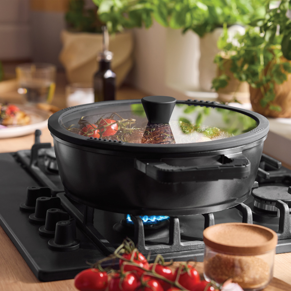 Crna šerpa sa providnim poklopcem na plinskom šporetu, sa paradajzom i začinima.