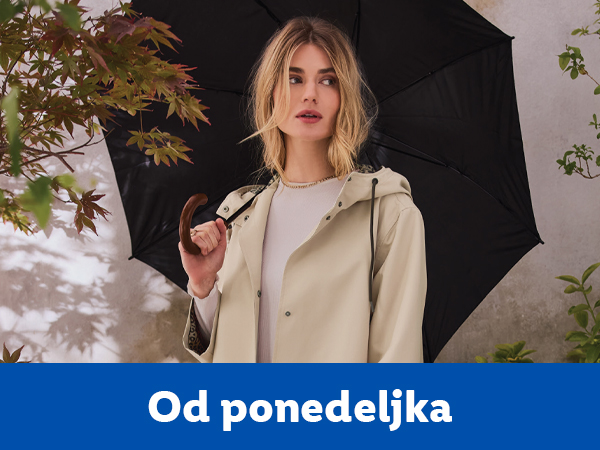 Žena u bež kabanici drži crni kišobran, sa zelenim lišćem u pozadini.
