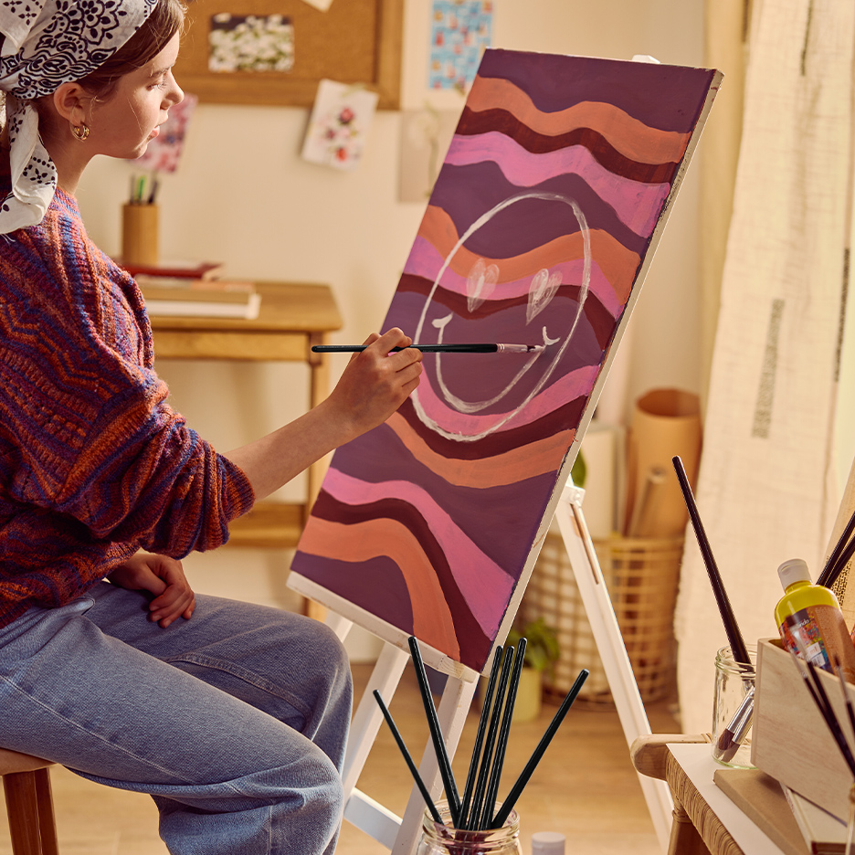 Devojka slika smajlija na platnu sa talasastim prugama, koristeći četkicu i boje.