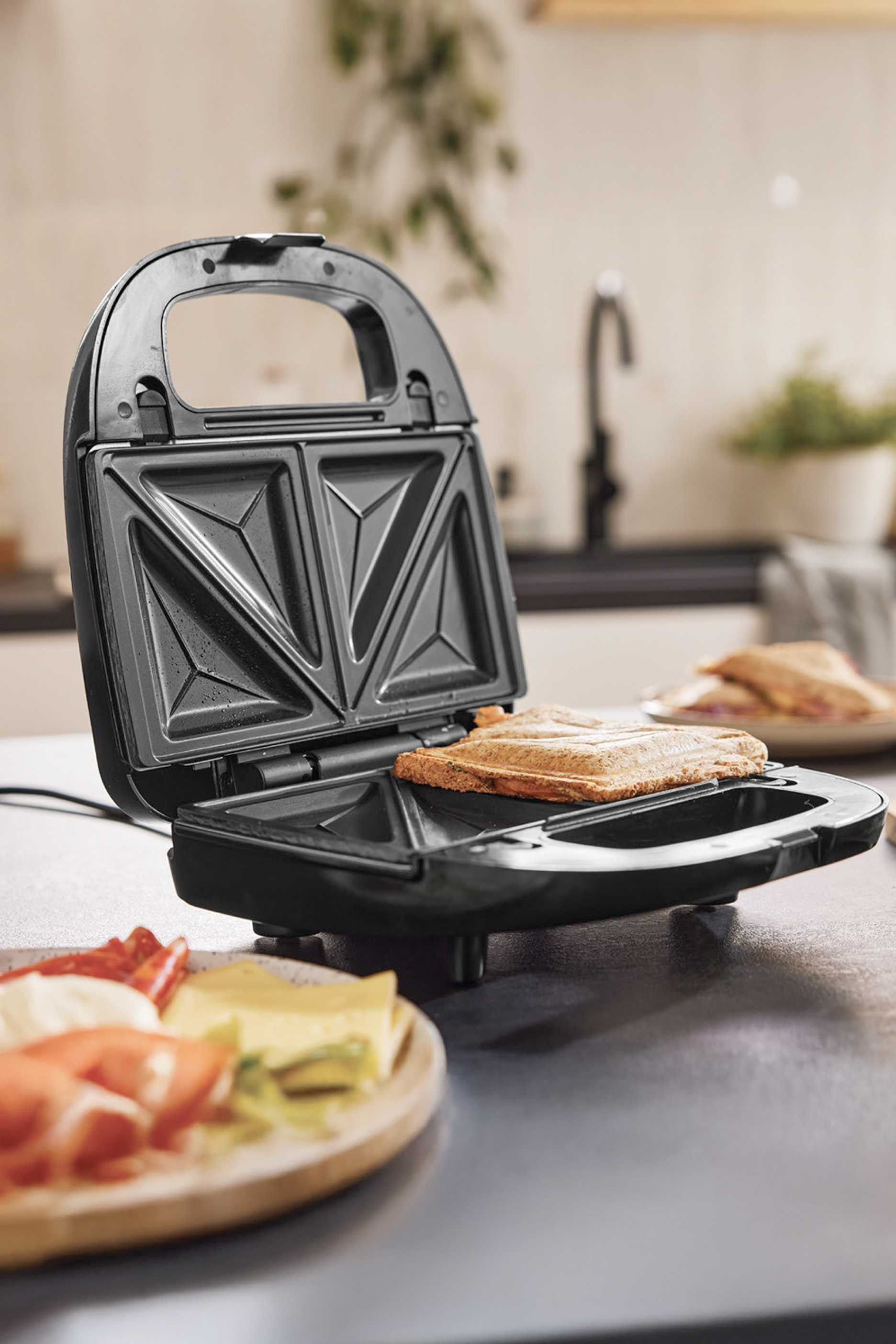 Aparat za sendviče sa tostiranim sendvičem i tanjirom sa sirom i mesom.