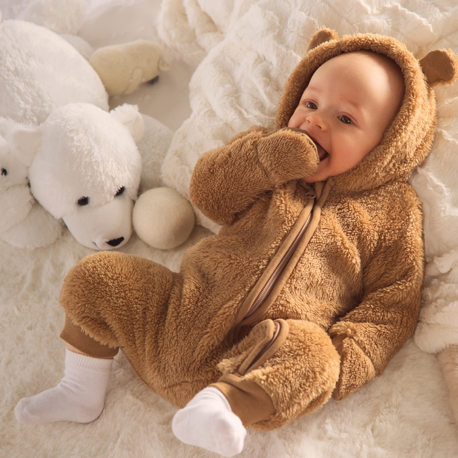 Beba u plišanom meda odelu, okružena belim plišanim igračkama.