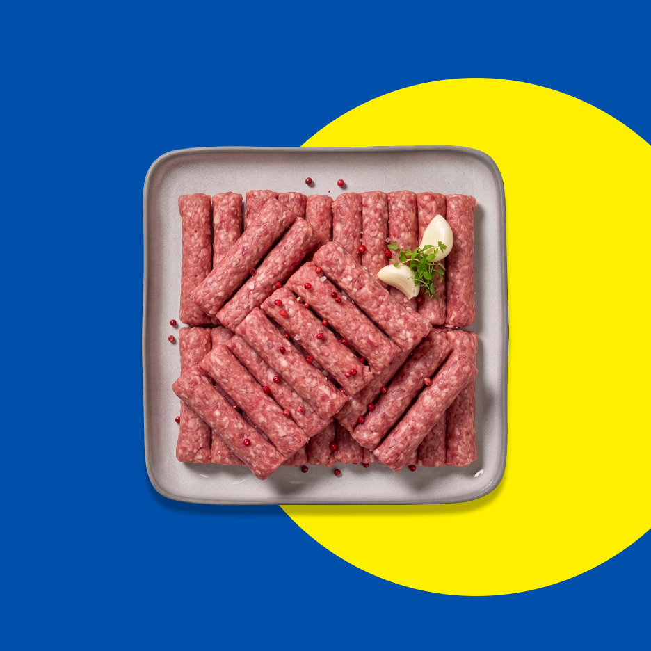 Sveži ćevapi servirani na sivoj tacni sa čenovima belog luka i začinskim biljem, na plavo-žutoj pozadini.