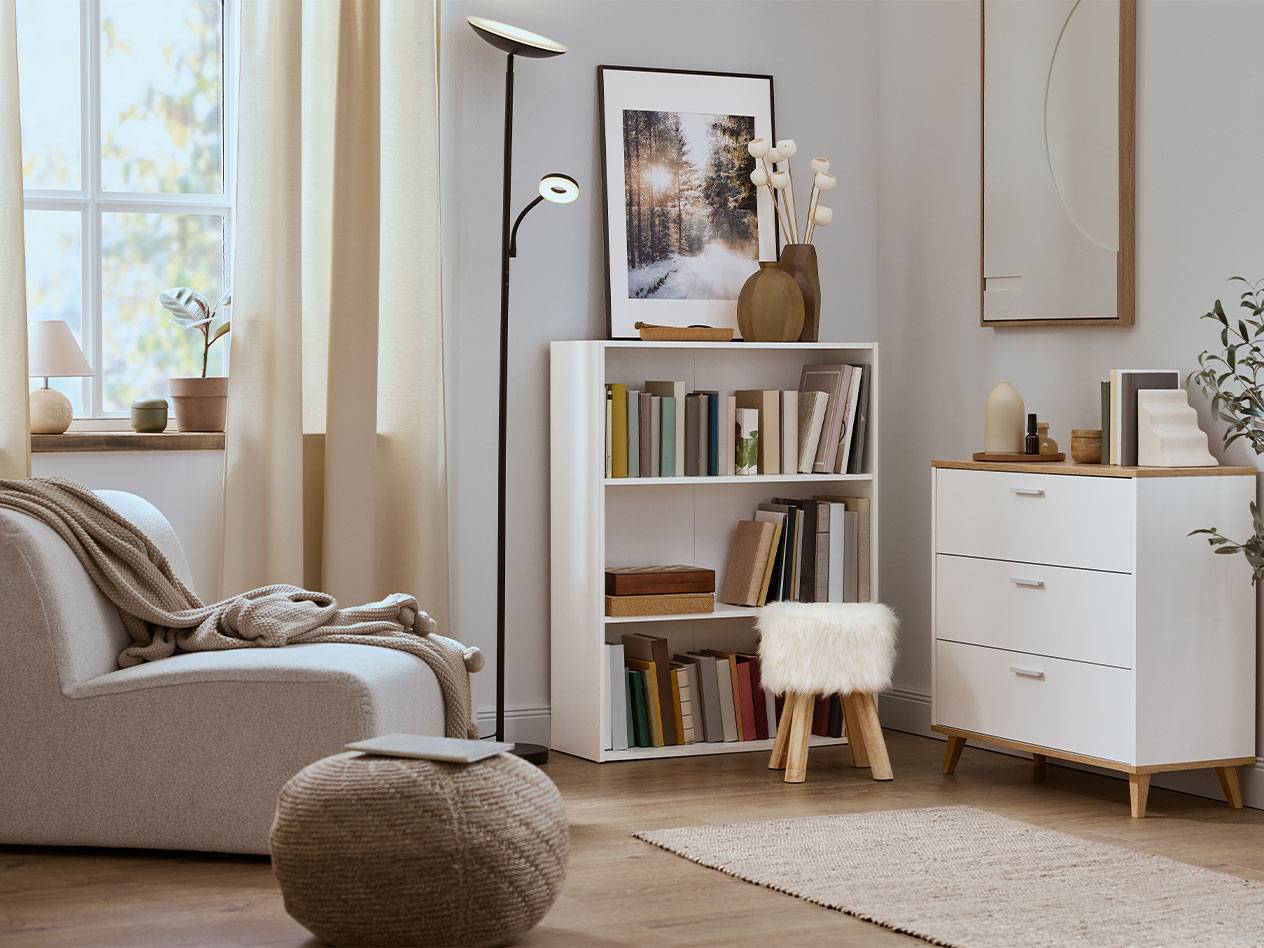 Udobna dnevna soba sa foteljom, policom za knjige i komodom u neutralnim bojama.