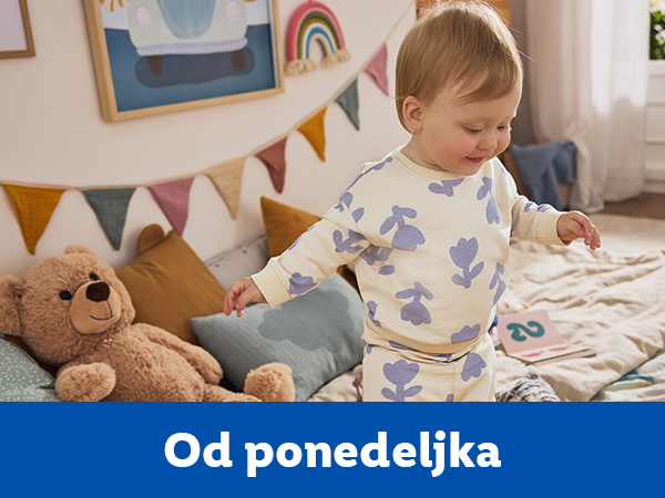 Beba u pidžami sa cvetnim dezenom stoji pored plišanog medveda i jastuka.