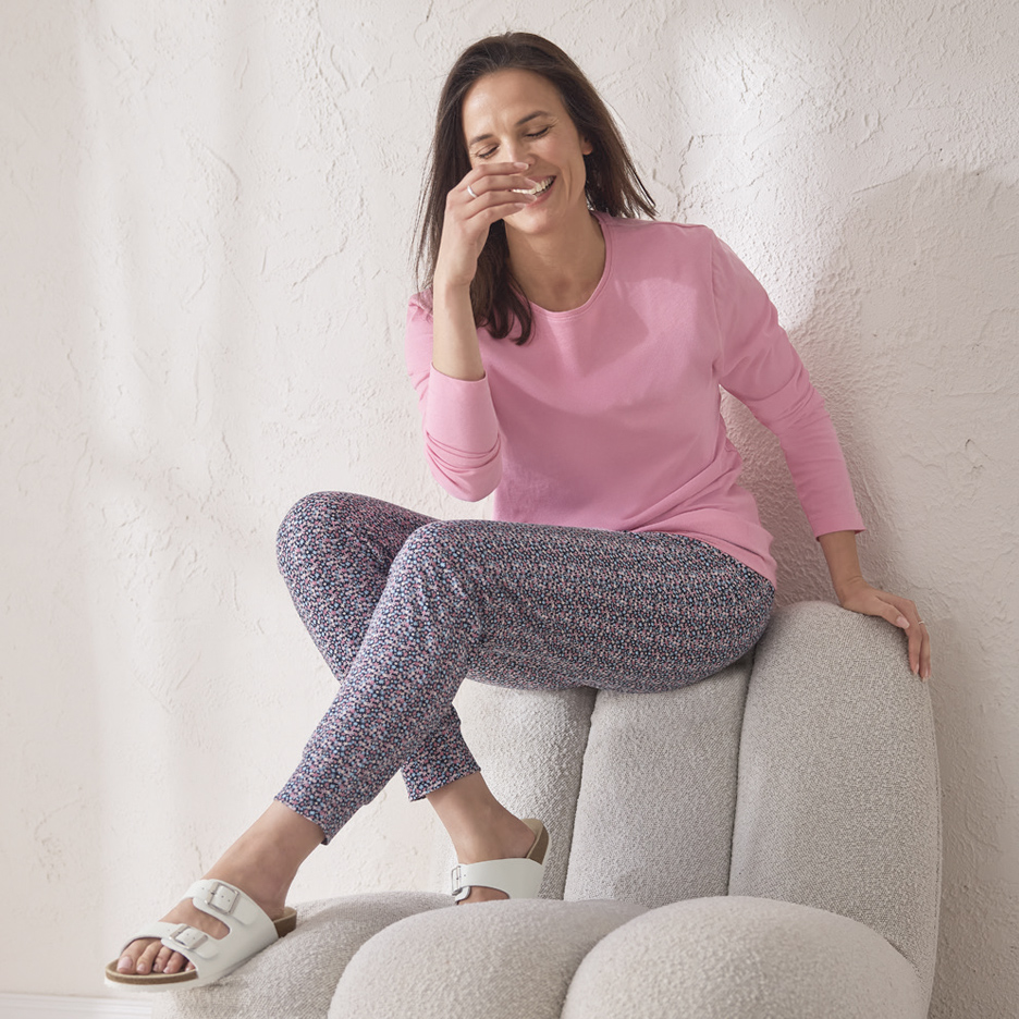 Žena u ružičastoj majici dugih rukava i cvetnim pantalonama sedi na stolici i smeje se.