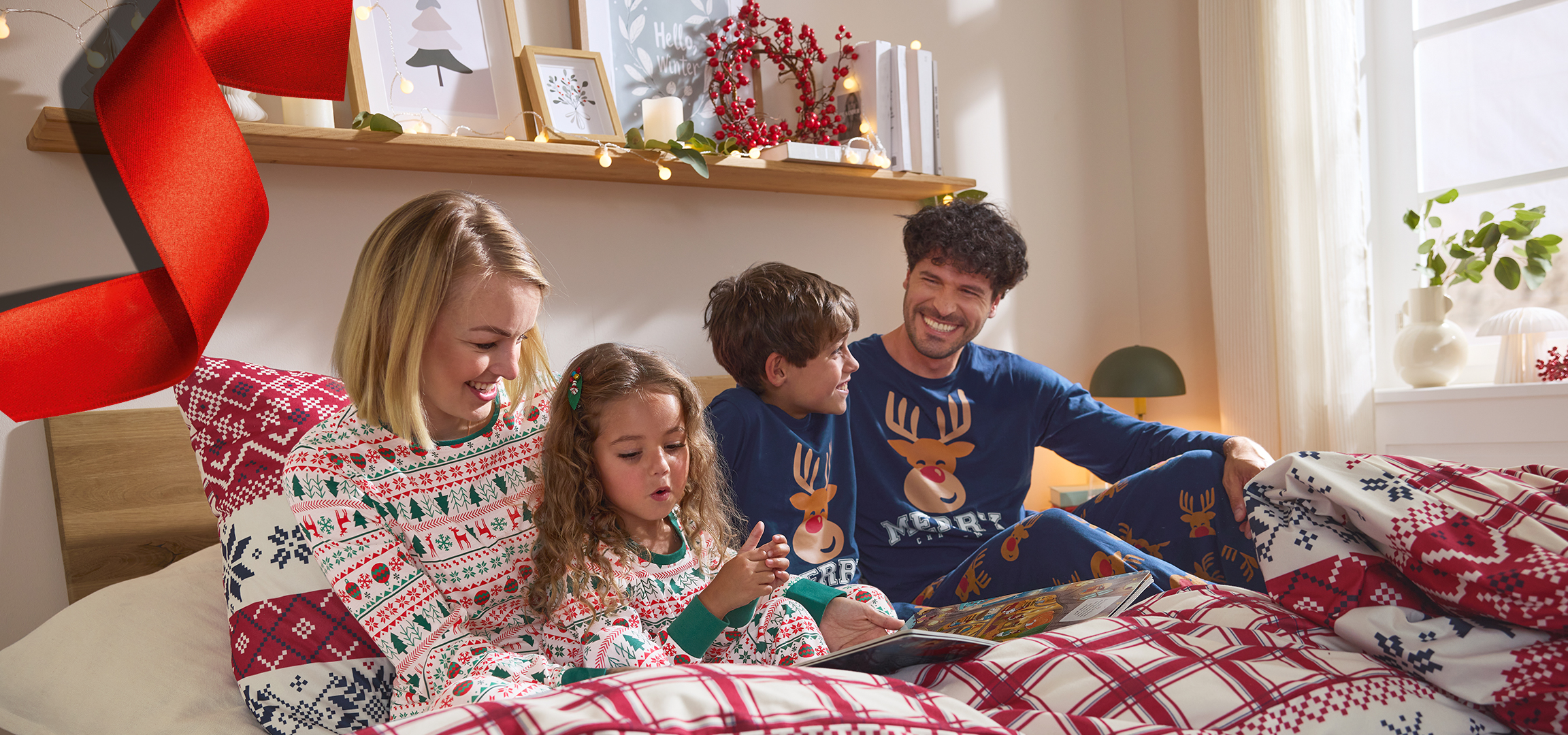 Porodica u božićnim pidžamama čita knjigu u krevetu, sa izloženim pidžamama sa strane.