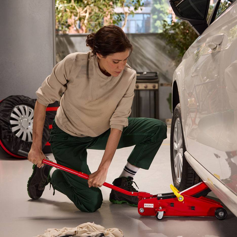 Žena koristi crvenu hidrauličnu dizalicu za podizanje belog automobila, sa rezervnim točkom u blizini.