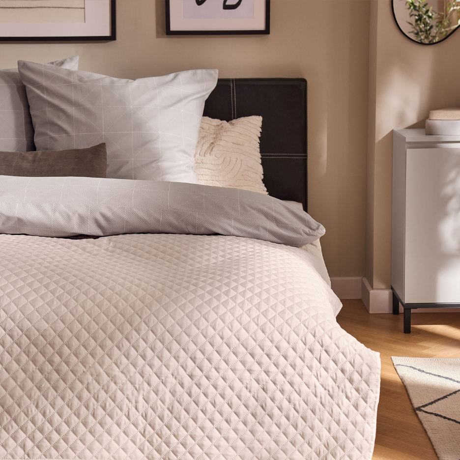 Krevet sa sivom posteljinom, bež prekrivačem i tamnim uzglavljem u spavaćoj sobi.