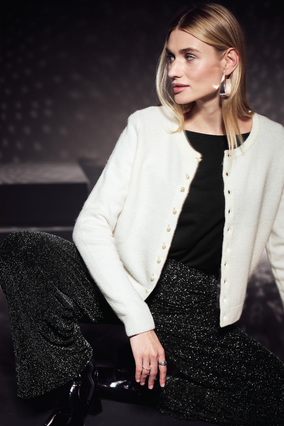Žena u belom kardiganu sa perlama, crnoj majici i svetlucavim pantalonama.