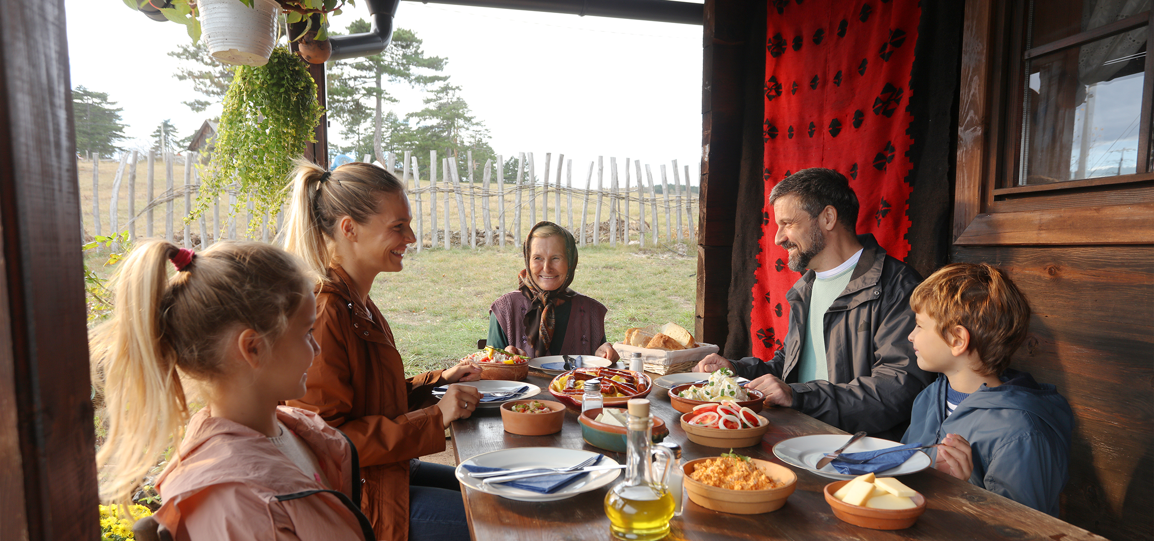 Porodica uživa u obroku na otvorenom, dok se na desnoj strani nalaze činije sa bobičastim voćem i sirom.