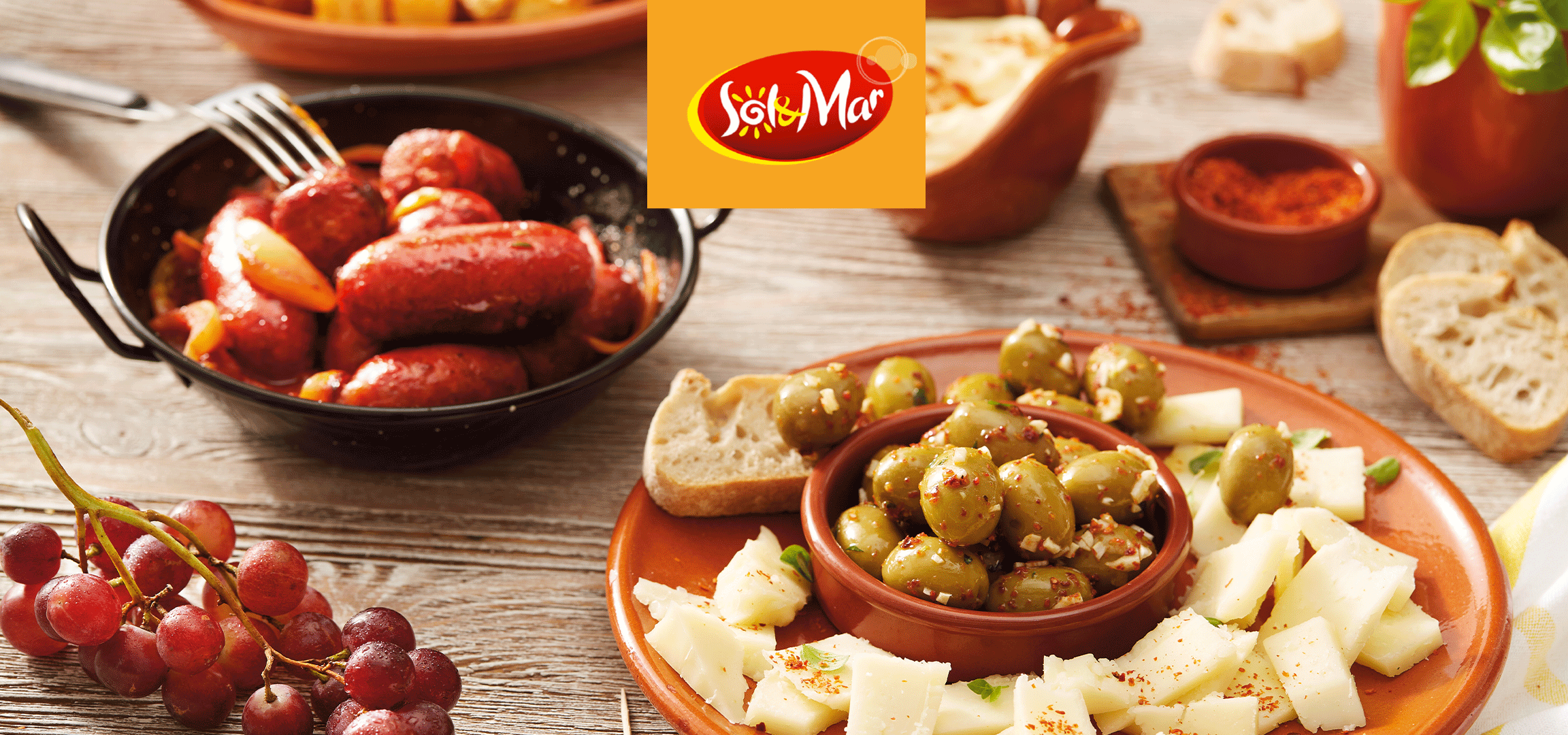 Masline, sir i grožđe na drvenom stolu, sa logotipom u uglu.