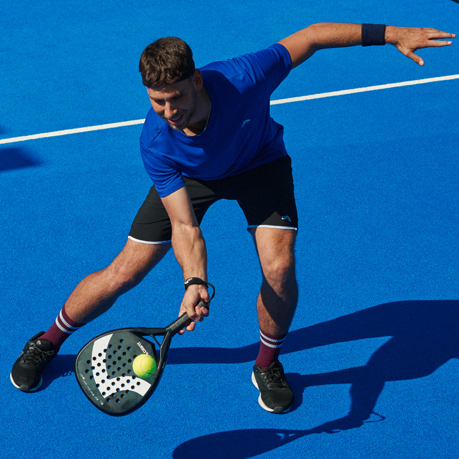 Muškarac u plavoj majici i crnom šortsu igra padel sa reketom i lopticom.