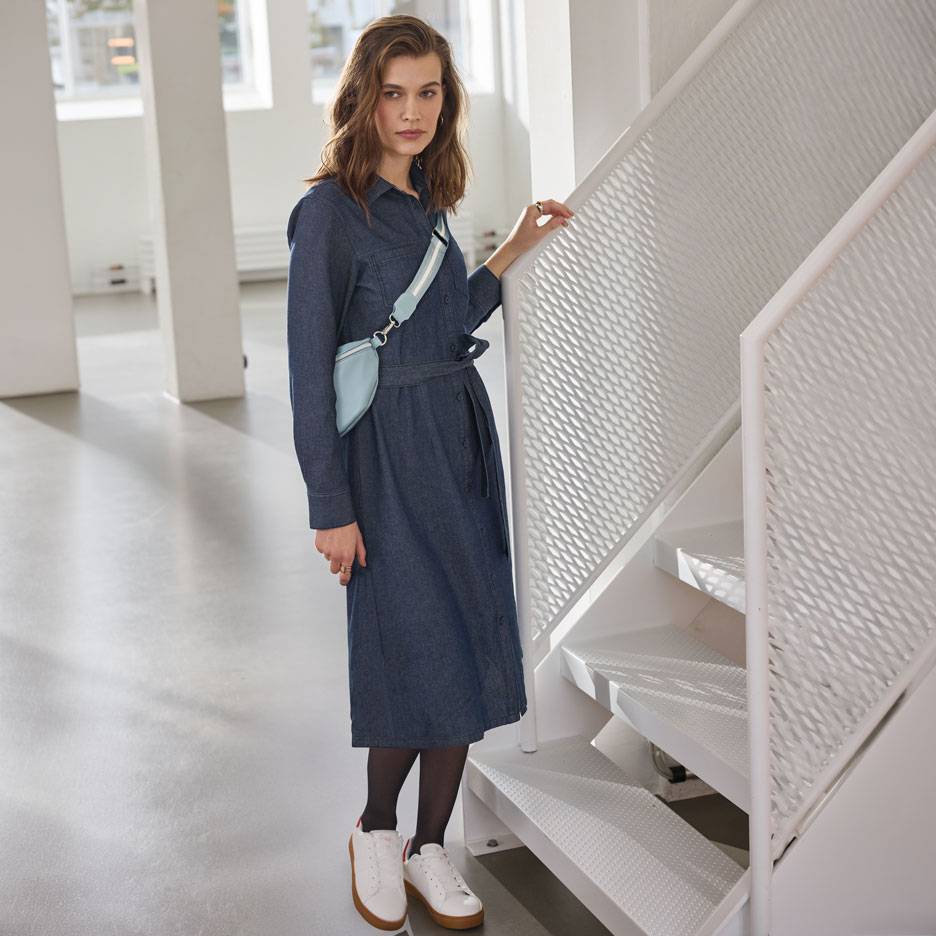 Žena u teksas košulja-haljini, crnim hulahopkama i belim patikama, sa svetloplavom torbicom oko struka.