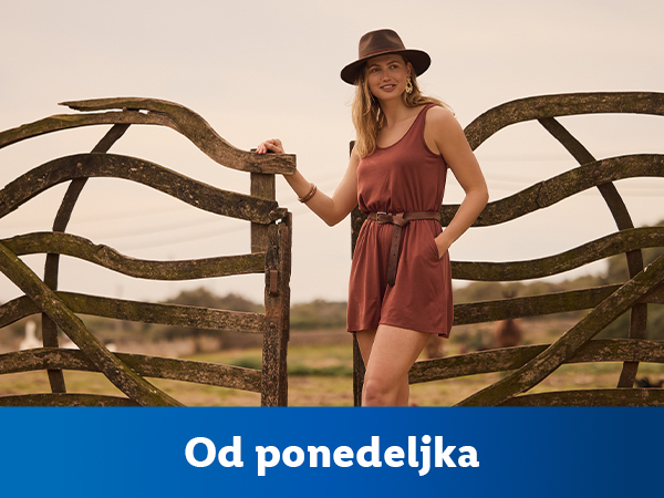 Žena u smeđoj haljini i šeširu stoji pored drvene ograde. Od ponedeljka.