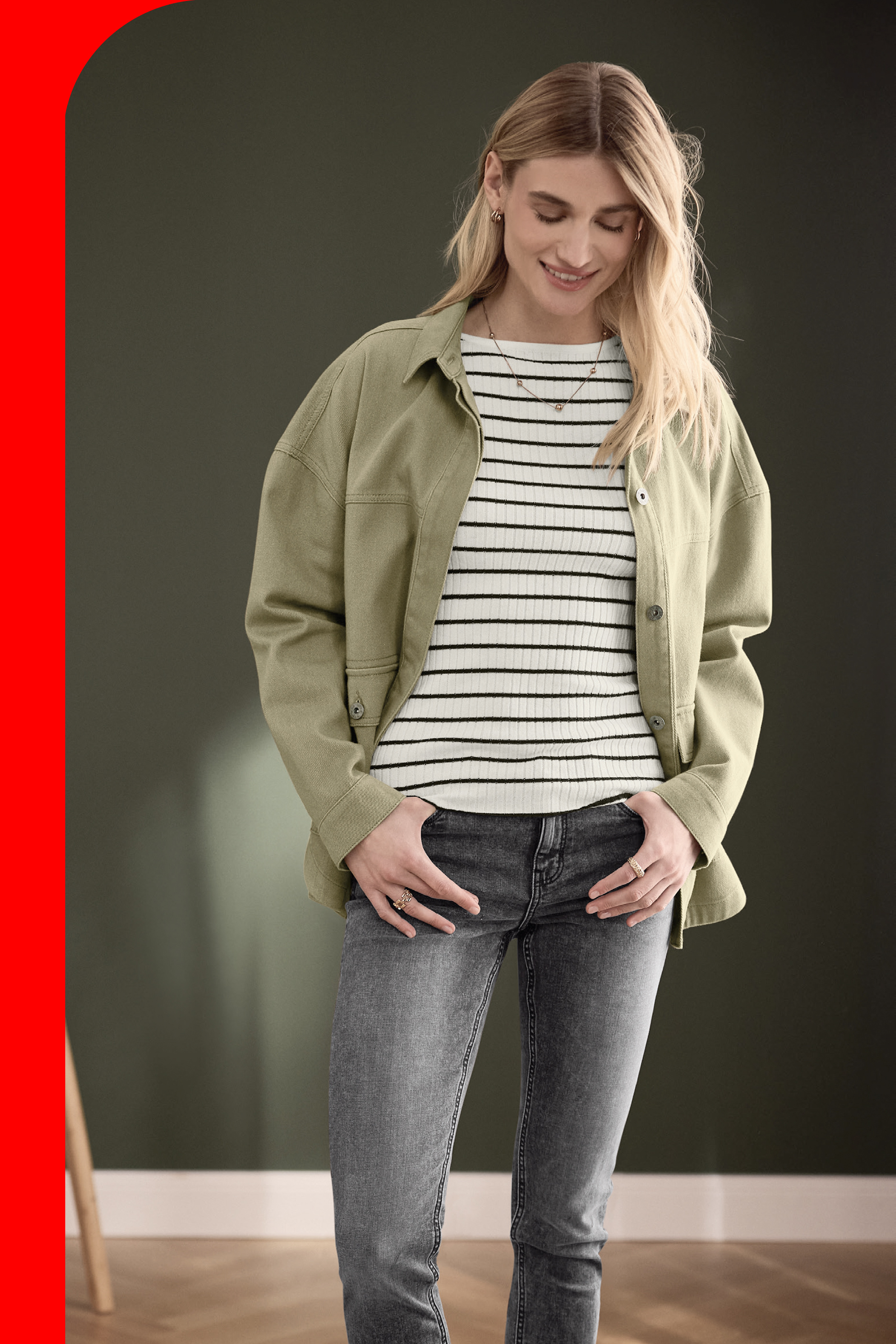Žena u maslinasto zelenoj jakni, prugastoj majici i sivim farmerkama, sa rukama u džepovima.