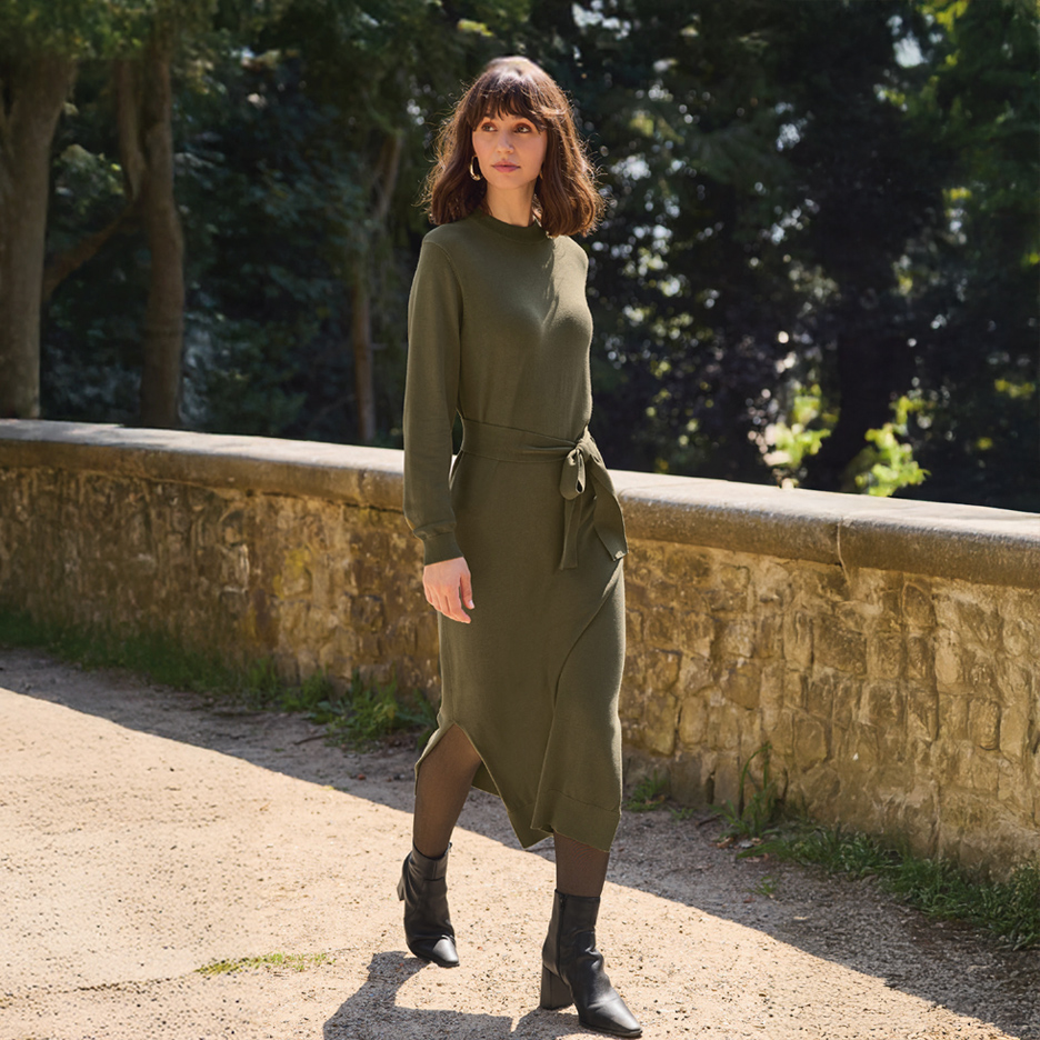 Žena u maslinastozelenoj pletenoj haljini sa kaišem i crnim čizmama hoda stazom.