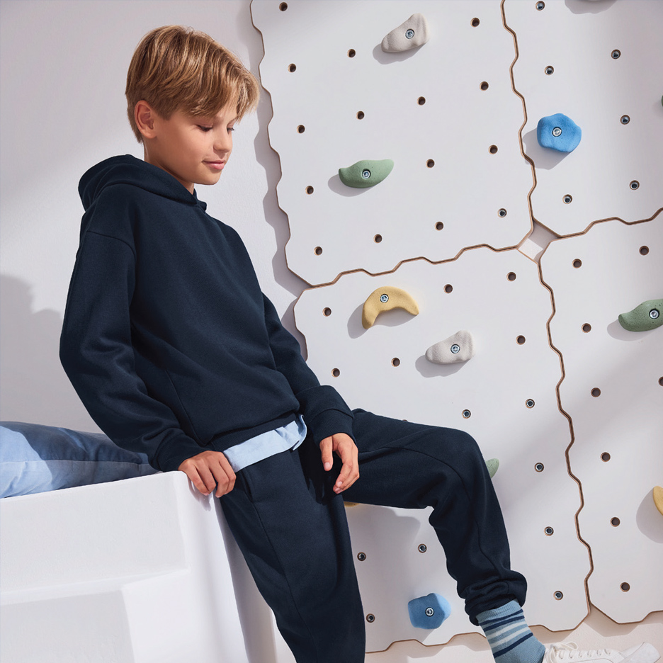 Dečak u tamnoplavoj dukserici sedi pored zida za penjanje sa šarenim držačima.