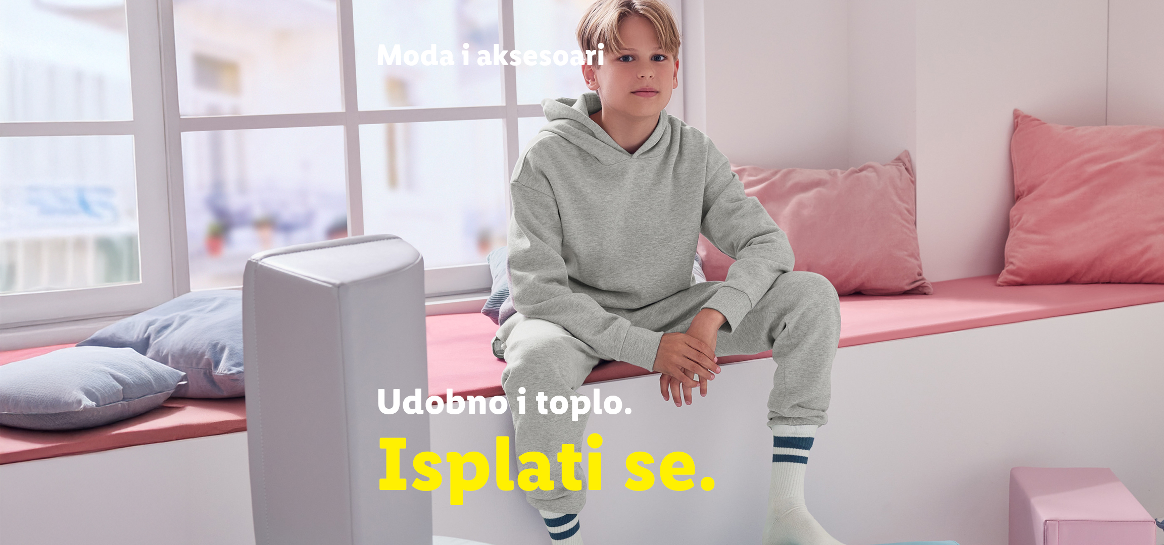 Dečak u sivom duksu i trenerci, sa ružičastim i plavim jastucima, pored ružičaste i tamnoplave trenerke.