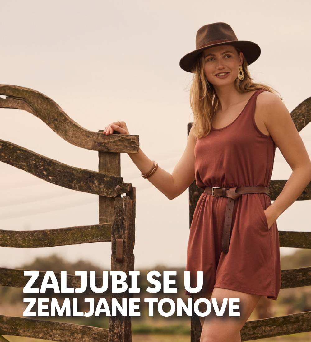 Žena u smeđoj haljini i šeširu stoji pored drvene ograde.
